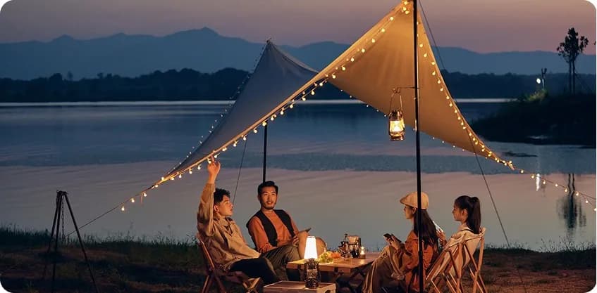 Friends camping by a lakeside under string lights at dusk