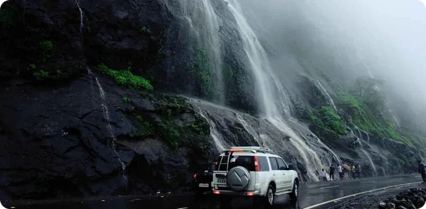 Self-drive SUV parked near misty mountain waterfalls on a monsoon road