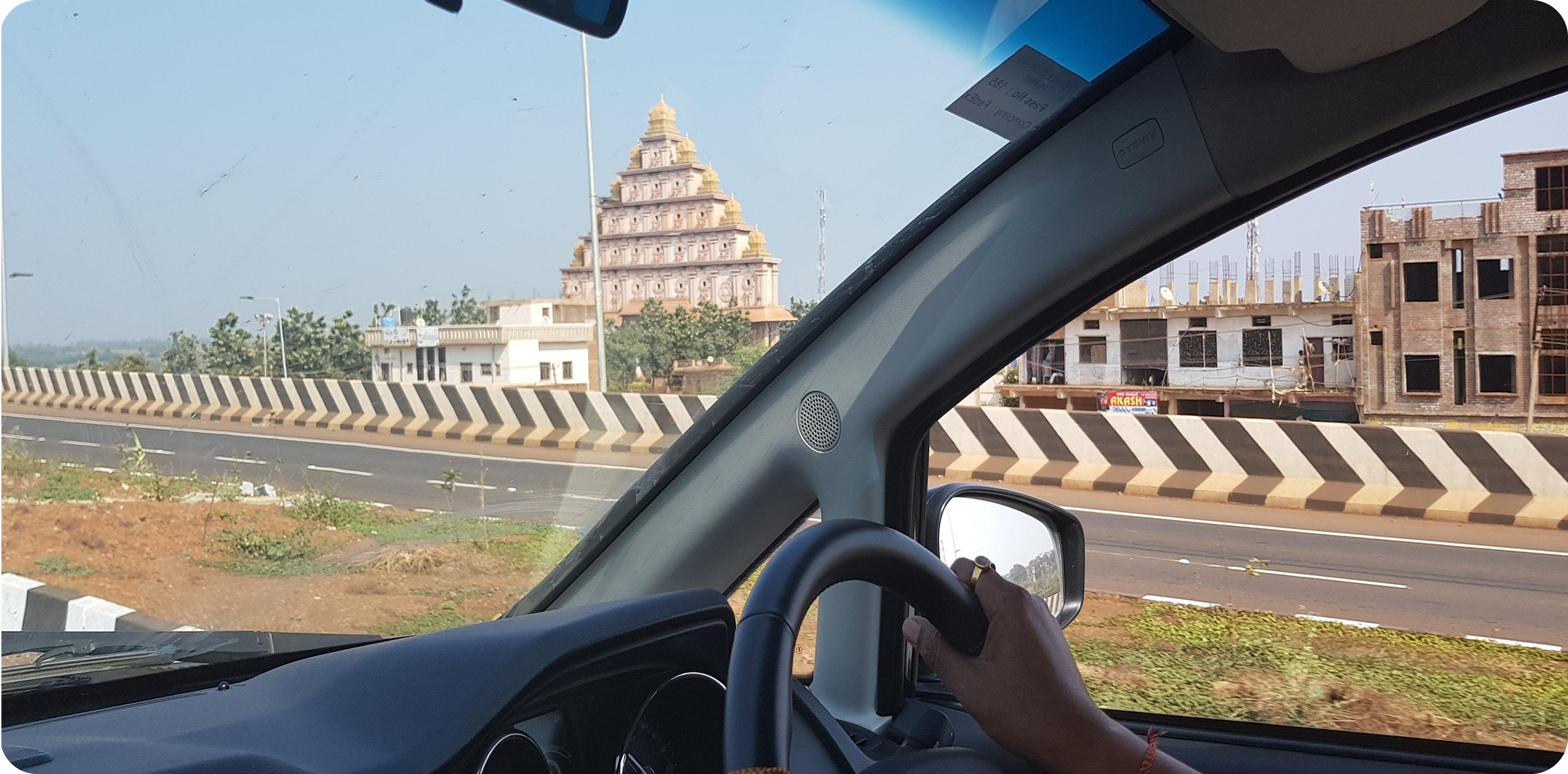 The image appears to show a view from inside a car driving past a multi-tiered, pyramid-shaped structure resembling a large meditation or spiritual monument next to a highway in India.
